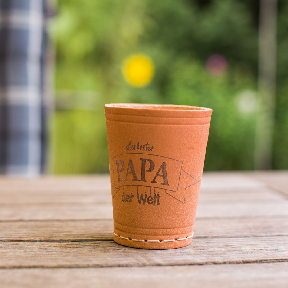 Würfelbecher mit Gravur Motiv "Allerbester Papa der Welt" auf Tisch draußen stehend im Hintergrund leicht angedeutet eine Gartenanlage.