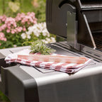 Braune Buchenholz-Grillzange mit individueller Lasergravur „Grillprofi Sebastian“, liegend auf kariertem Geschirrtuch neben Grill und Kräutern.